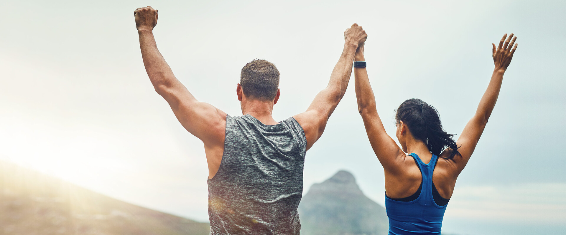 Ein sportliches Paar vor alpiner Kulisse reckt triumphierend die Arme in den Himmel. Das Bild illustriert das Thema Körperpflege für einen fitten Körper. 