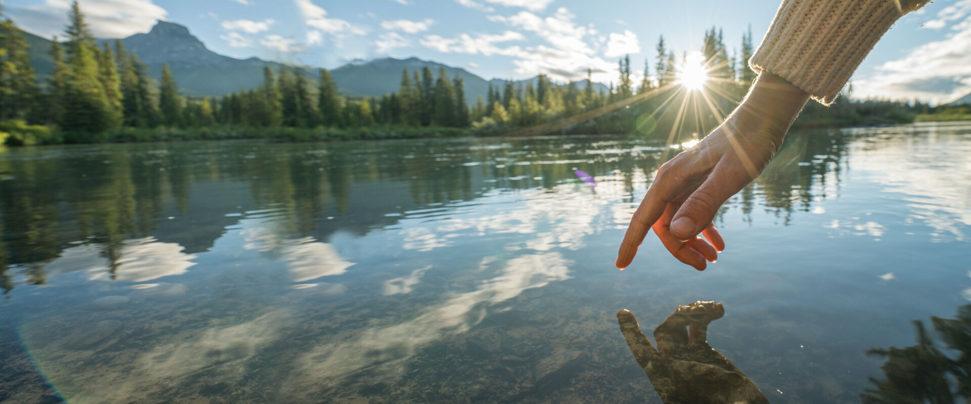 Ein Finger schwebt direkt über einem Bergsee und berührt fast die Oberfläche. Das Bild leitet das Thema Nagelpflege ein.