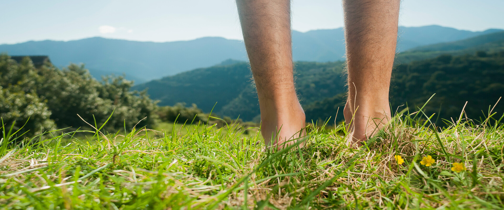 Zwei männliche, nackte Beine stehen vor einem Alpenpanorama im Gras und symbolisieren das Thema Blasen an den Füßen.